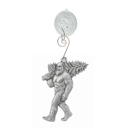 A silver metal hanging ornament featuring a detailed Bigfoot figure carrying pine branches on his back, suspended from a clear circular suction cup with a wire loop, shown against a plain white background.