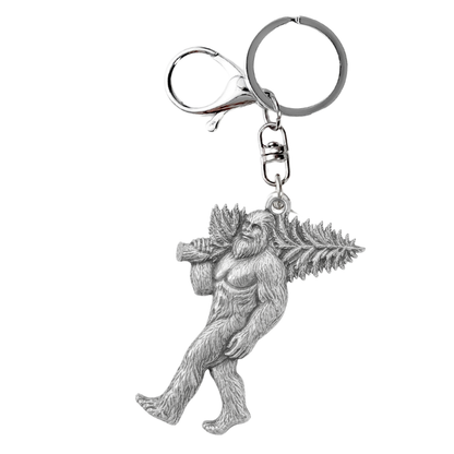 A silver metal keychain featuring a detailed figurine of a Bigfoot carrying a pine tree, attached to a classic split ring and lobster clasp, shown on a plain white background.