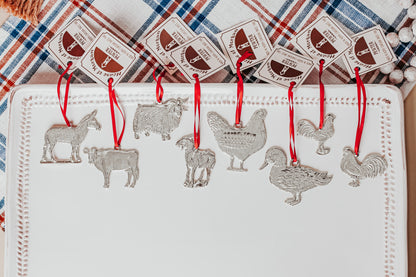 A set of pewter farm animal ornaments with red ribbons, including designs of a cow, chicken, horse, goat, sheep, donkey, ram, and rooster, displayed on a white textured surface with a plaid background.