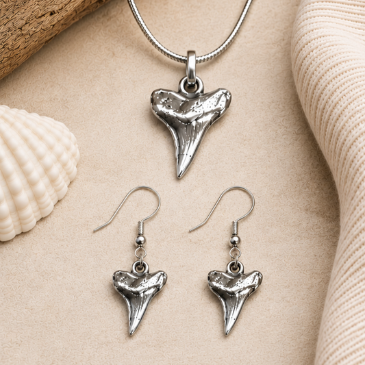 Silver shark tooth necklace and earrings on a sandy surface with shells.